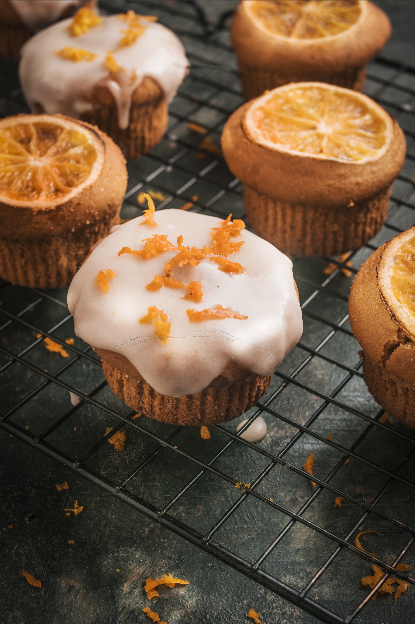 Close-up of iced blood orange cupcakes on a cooling rack, finished with a thin white glaze and scattered fresh orange zest.